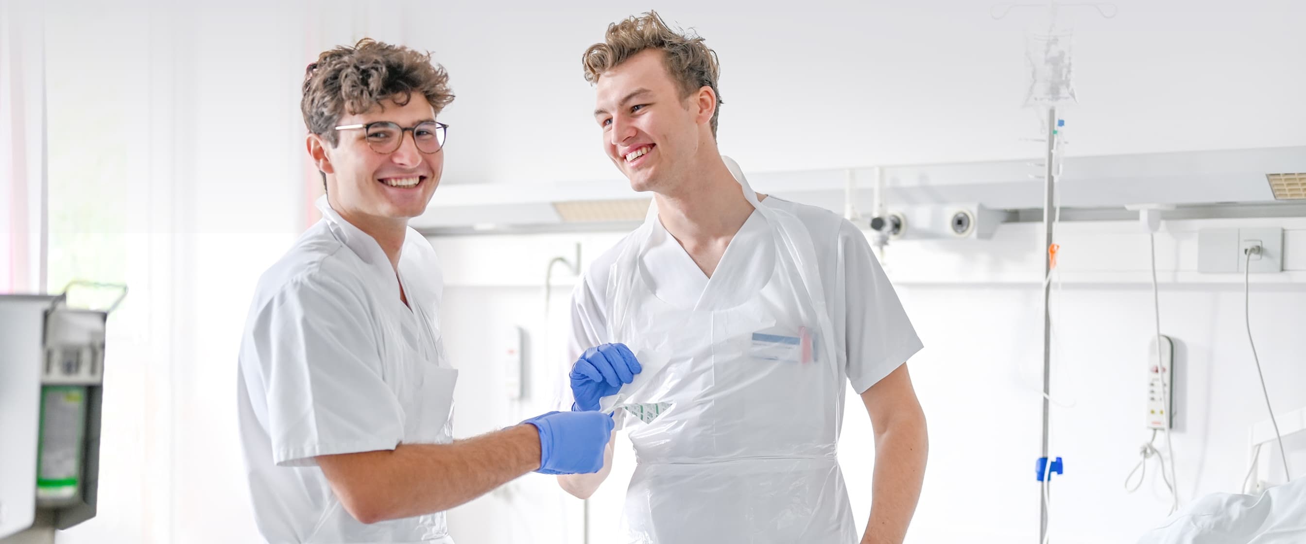 Zwei Pflegeazubis stehen in einem Patientenzimmer, einer davon blickt in die Kamera