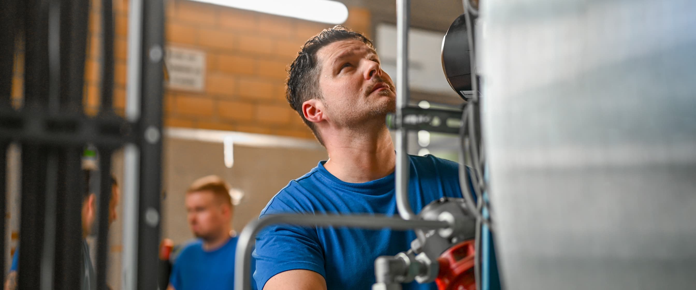 Ein Elektroniker prüft eine Maschine im Keller des TUM Klinikums. Zwei Kollegen stehen im Hintergrund und besprechen etwas.