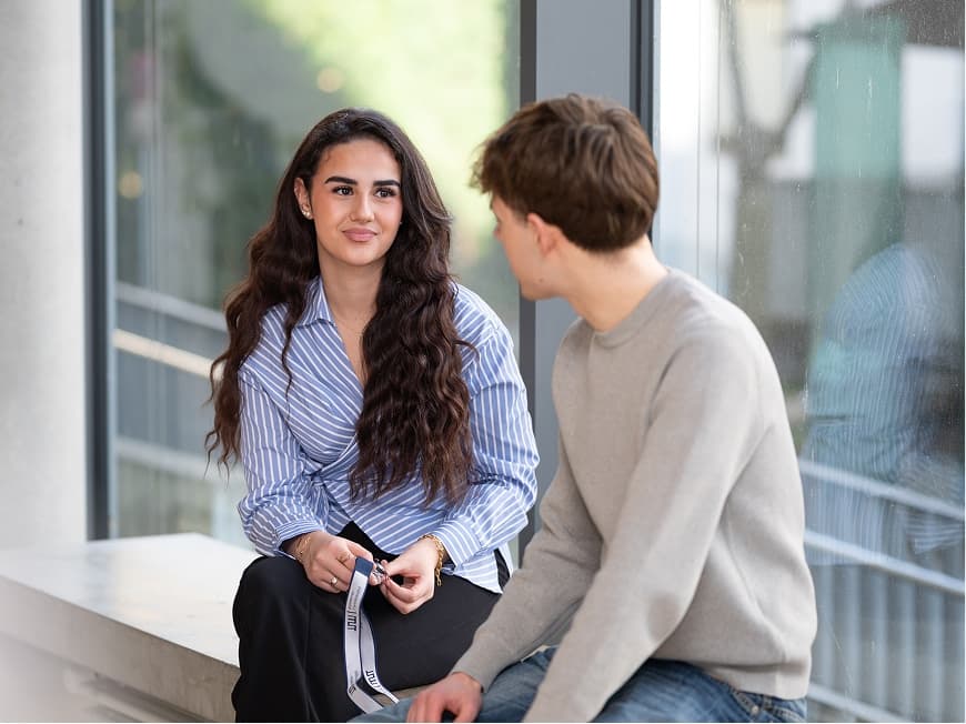 Eine Auszubildende und ein Auszubildender zur Kauffrau bzw. zum Kaufmann im Gesundheitswesen sitzen im Krankenhausflur auf einer Fensterbank und tauschen sich zu einem aktuellen Ausbildungsthema aus
