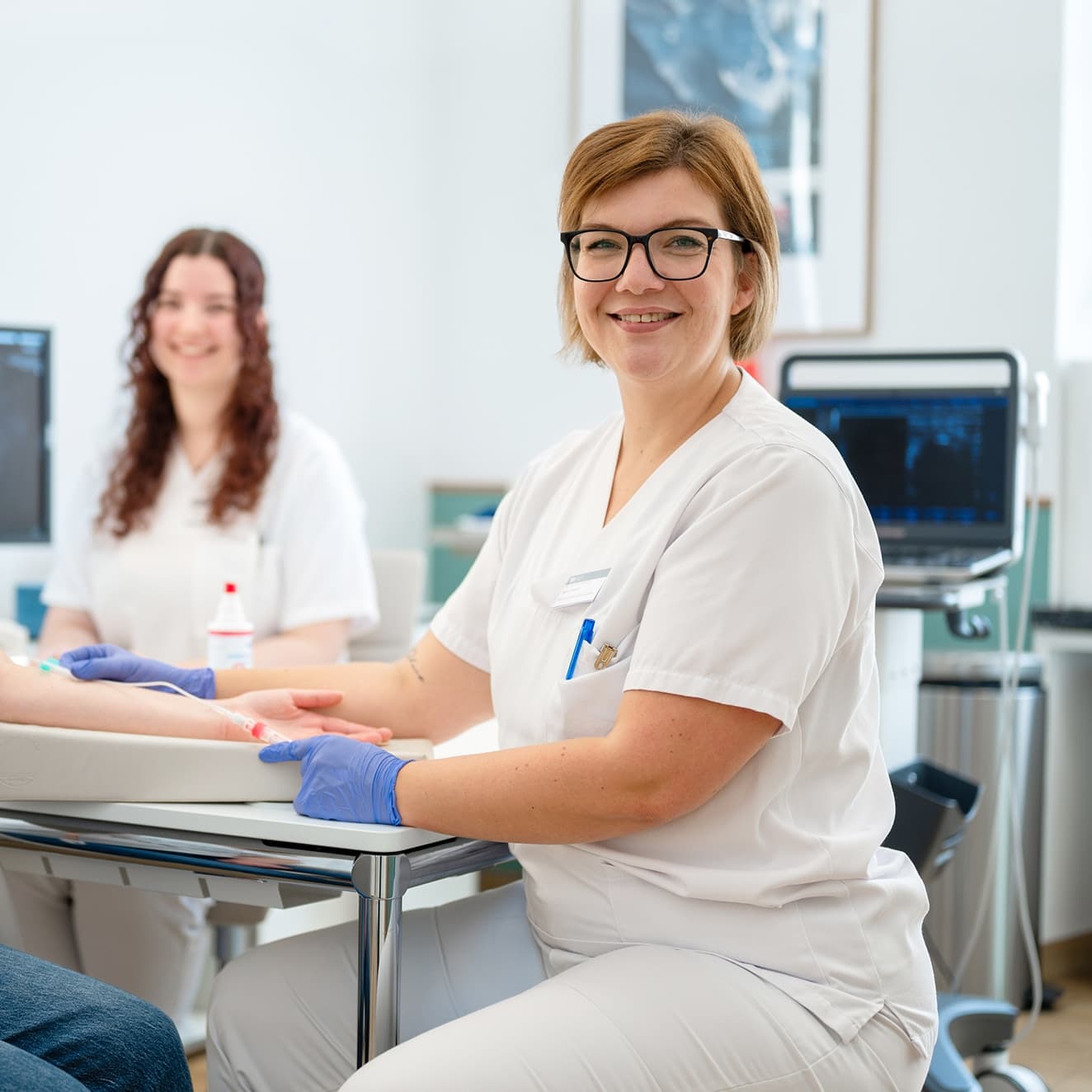 Eine medizinische Fachangestellte nimmt einer Patientin Blut ab während eine Kollegin am Rechner die Patientenakte aktualisiert