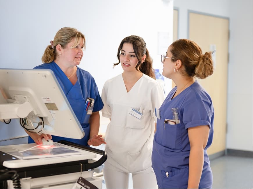 Zwei Auszubildende zur Pflegefachfrau bekommen von einer Praxisanleiterin etwas erklärt