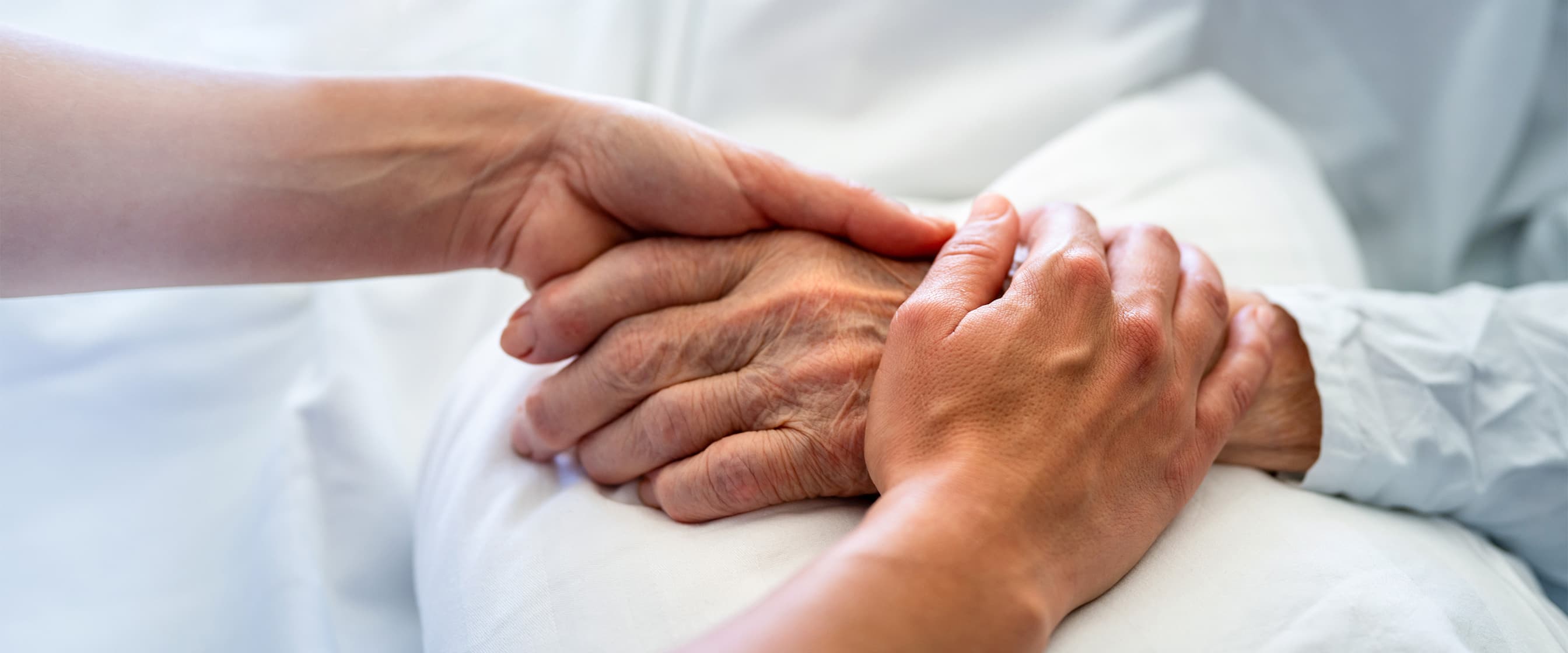 Die Hände einer Pflegefachkraft halten die Hände einer älteren Palliativpatientin, die im Krankenbett liegt