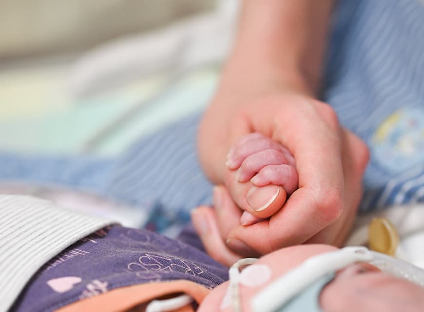 Die Hand einer Pflegefachkraft hält den Fuß eines Neugeborenen, das in einem Inkubator auf der Neonatologie liegt