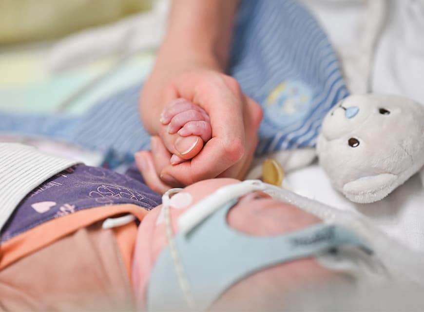 Die Hand einer Pflegefachkraft hält den Fuß eines Neugeborenen, das in einem Inkubator auf der Neonatologie liegt
