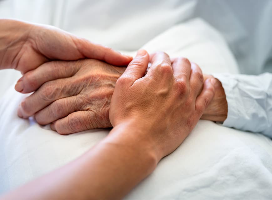 Die Hände einer Pflegefachkraft halten die Hände einer älteren Palliativpatientin, die im Krankenbett liegt